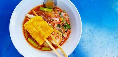 Close up Spicy Pink seafood flat noodles soup with chopsticks, deep fried pork wonton in white bowl on blue table or background. photo