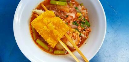 Close up Spicy Pink seafood flat noodles soup with chopsticks, deep fried pork wonton in white bowl on blue table or background. photo