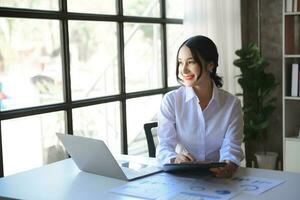 Beautiful Asian woman using laptop and tablet while sitting at her working place. Concentrated at work. photo