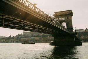 turista barco navegación debajo el cadena puente terminado el río Danubio en Budapest con el Dom brillante detrás durante el puesta de sol en dorado hora foto