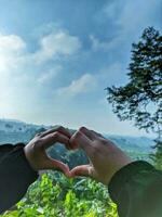 Hands of a woman in the shape of a heart with a finger and green nature background. A love concept with a sky background and space for text. Feelings and emotions concept. photo