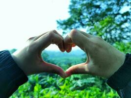 Hands of a woman in the shape of a heart with a finger and green nature background. A love concept with a sky background and space for text. Feelings and emotions concept. photo