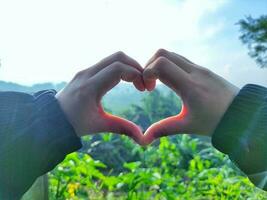 Hands of a woman in the shape of a heart with a finger and green nature background. A love concept with a sky background and space for text. Feelings and emotions concept. photo