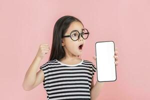 asiático pequeño niña participación teléfono inteligente Bosquejo de blanco pantalla y sonriente en blanco antecedentes. foto