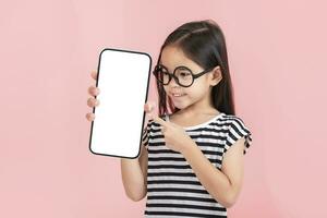 asiático pequeño niña participación teléfono inteligente Bosquejo de blanco pantalla y sonriente en blanco antecedentes. foto