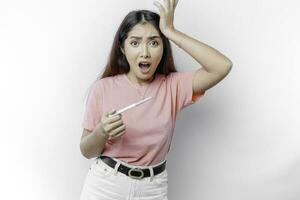 Unhappy woman wearing pink t-shirt looking at pregnancy test alone, confused young female shocked by result, bad news, covering open mouth with hand, unwanted pregnancy concept photo