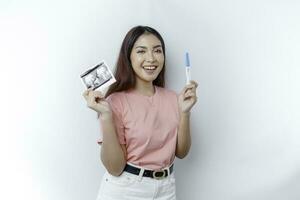 Happy young woman wearing pink t-shirt showing her pregnancy test and ultrasound picture, isolated on white background, pregnancy concept photo