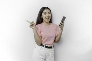 A portrait of a happy Asian woman wearing pink t-shirt, holding her phone, and pointing copy space on beside her, isolated by white background photo