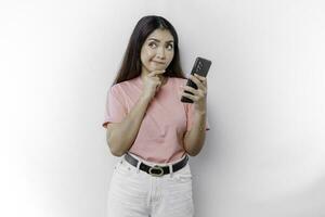A thoughtful young woman wearing pink t-shirt, holding her chin and her phone isolated by white background photo