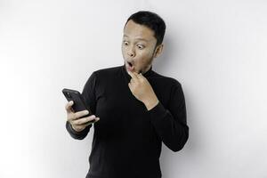 Shocked Asian man wearing black shirt holding his phone with his mouth wide open, isolated by white background photo