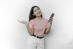 A portrait of a happy Asian woman wearing pink t-shirt, holding her phone, and pointing copy space on beside her, isolated by white background photo