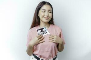 Happy young woman wearing pink t-shirt showing her pregnancy test and ultrasound picture, isolated on white background, pregnancy concept photo