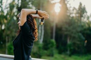 Profile shot of active motivated young woman dressed in black t shirt, raises dumbbell over head, poses against trees with sunset, has workout outdoor, wants to have muscular arms. Sport concept photo