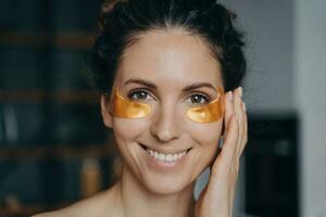 Smiling latina woman applying moisturizing golden under eye patches at home. Skincare beauty routine photo