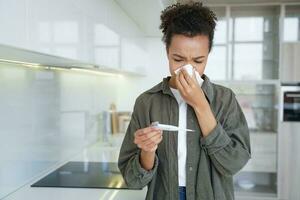 Sick mixed race girl blowing nose checks temperature using electronic thermometer at home. Flu fever photo