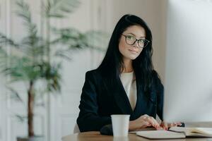 Serious successful female office worker in formal outfit, wears eyeglasses, busy with online work, poses at desktop, works on computer, reads business new or watches tutorial, enjoys working process. photo