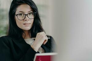 Self confident serious adult female employee looks though transparent glasses, writes information while working on project, makes plans, dressed in formal wear, notes some records in notepad photo