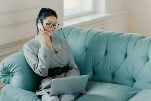 Pleased businesswoman in elegant clothes discusses financial issues with partner via mobile phone, poses on comfortable sofa, sits at modern laptop computer in home office. Technology, work concept photo