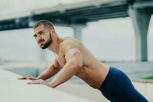 Bearded man does push-ups outdoors, warms up, poses near a bridge, has early morning workout. photo