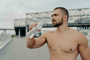 Muscular guy takes a break, drinks water, cares about hydration, poses outside after jogging. photo