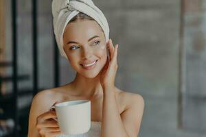 Pretty woman applies face cream, minimal makeup, wrapped in bath towel, enjoys coffee at home. Beauty, relaxation concept. photo