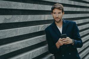 Thoughtful young bearded man standing outside holding mobile, typing on phone keyboard photo