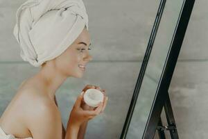 Happy woman with bare shoulders, towel on head, applies face cream, admires reflection. Self-care. photo