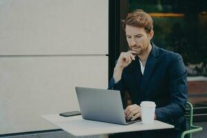Handsome pensive young man browsing web on laptop while working remotely in sidewalk cafe photo