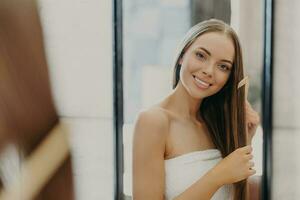 hermosa mujer sonrisas y peines su Derecho cabello, envuelto en toalla, en baño. De las mujeres belleza concepto foto