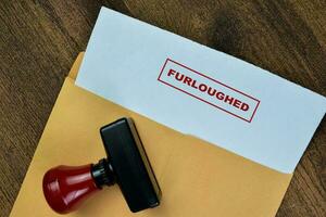 Concept of Red Handle Rubber Stamper and Furloughed text above brown envelope isolated on on Wooden Table. photo