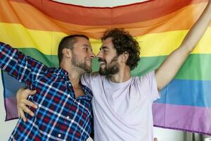 Two young gay lovers kissing each other affectionately. Two young male lovers standing together against a pride flag. Affectionate young gay couple sharing a romantic moment together. photo