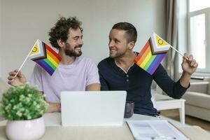Male lovers video calling their friends in their living room at home. Young gay couple smiling cheerfully while greeting their friends on a video call. Holding LGBTQ flags and celebrating pride month photo