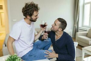 lgbtq Pareja abrazando cada otro y hundiéndose vino adentro. dos romántico joven masculino amantes mirando a cada otro mientras sentado juntos en su vivo habitación. joven gay Pareja siendo romántico a hogar. foto