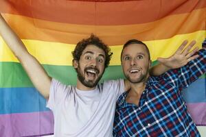 dos joven gay amantes mirando a cada otro afectuosamente. dos joven masculino amantes en pie juntos en contra un orgullo bandera. cariñoso joven gay Pareja compartiendo un romántico momento juntos. foto