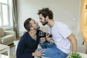 lgbtq Pareja abrazando cada otro y Bebiendo vino adentro. dos romántico joven masculino amantes mirando a cada otro mientras sentado juntos en su vivo habitación. joven gay Pareja siendo romántico a hogar. foto