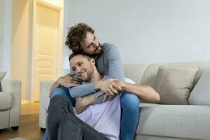 encantador gay Pareja besos mientras sentado en su vivo habitación a hogar. dos romántico joven masculino amantes teniendo divertido adentro. joven gay Pareja vivo juntos. foto