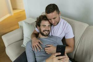 linda joven gay Pareja vídeo vocación su amigos en su vivo habitación a hogar. dos masculino amantes sonriente alegremente mientras saludo su amigos en un teléfono inteligente joven gay Pareja sentado juntos. foto