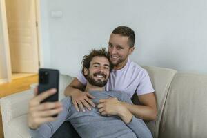 linda joven gay Pareja vídeo vocación su amigos en su vivo habitación a hogar. dos masculino amantes sonriente alegremente mientras saludo su amigos en un teléfono inteligente joven gay Pareja sentado juntos. foto