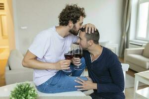 LGBTQ couple embracing each other and drinking wine indoors. Two romantic young male lovers looking at each other while sitting together in their living room. Young gay couple being romantic at home. photo