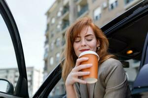 Happy young woman with coffee having a brake in her car. Side view of woman with coffee to go in hand. Young woman drinking coffee in her car photo