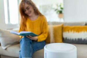 Modern air humidifier and blurred woman resting, reading a book on sofa on background photo