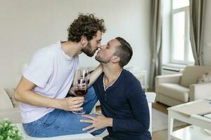 LGBTQ couple embracing each other and srinking wine indoors. Two romantic young male lovers looking at each other while sitting together in their living room. Young gay couple being romantic at home. photo