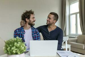 Lovely gay couple laughing together while sitting in their living room at home. Two romantic young male lovers having fun surfing the internet indoors. Young gay couple living together. photo