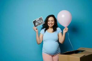 Excited happy pregnant woman smiles at camera, holds ultrasound scan her future child and pink balloon, blue background photo