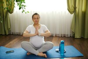 adulto embarazada mujer practicando embarazada yoga a hogar, sentado en azul aptitud estera, meditando en loto posición foto