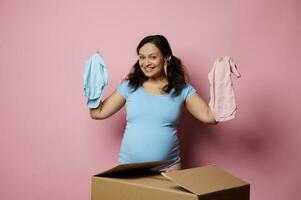 Beautiful happy pregnant brunette woman smiles looking at camera, holds newborn baby bodysuits, isolated pink background photo