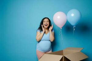 asombrado contento embarazada mujer expresando sorpresa a género revelar fiesta, esperando Gemelos bebé chico y chica, azul fondo foto