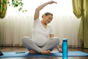 Positive happy pregnant woman practicing pregnancy yoga, outstretching right hand, sitting in lotus pose on exercise mat photo