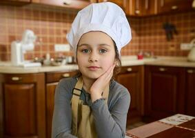 adorable niño niña en cocinero sombrero y delantal, pequeño panadero confitero mira a cámara. Cocinando clase. niños aprender culinario foto