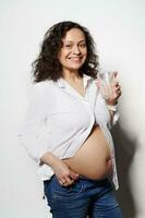Vertical portrait of happy smiling pregnant woman holding a glass of water, posing bare belly, white isolated background photo